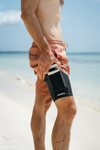 Shirtless man on a tropical beach inserting his phone into a black 800m thigh phone holder worn high on his right thigh.